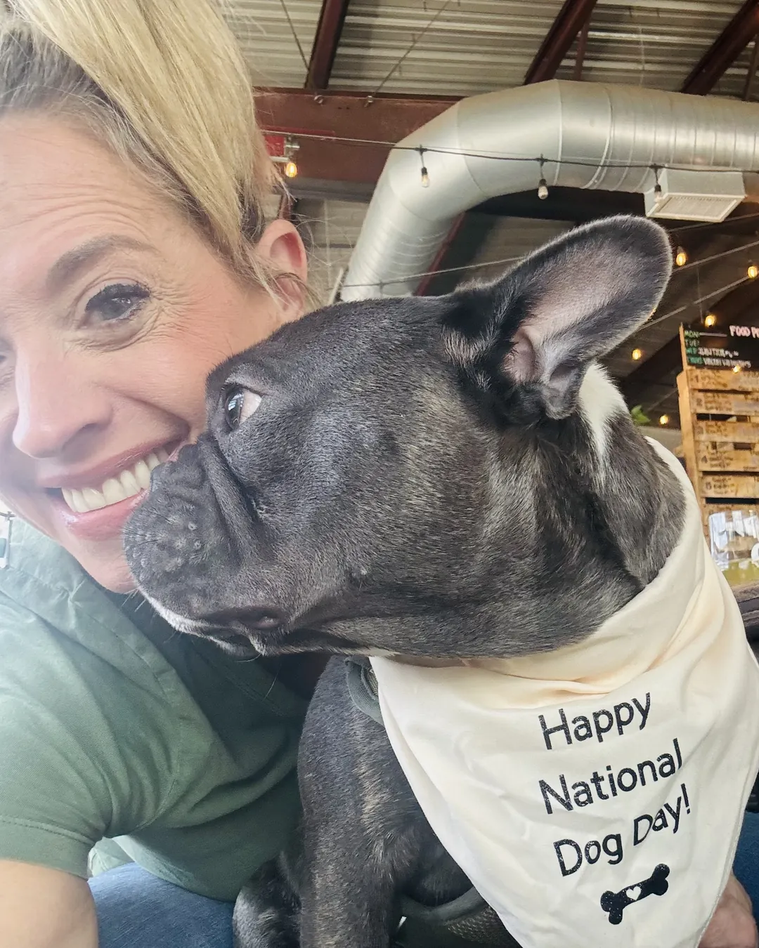 Gina Wilt smiling cheek to cheek with Dex, a dark French bulldog in a Happy National Dog Day bandana